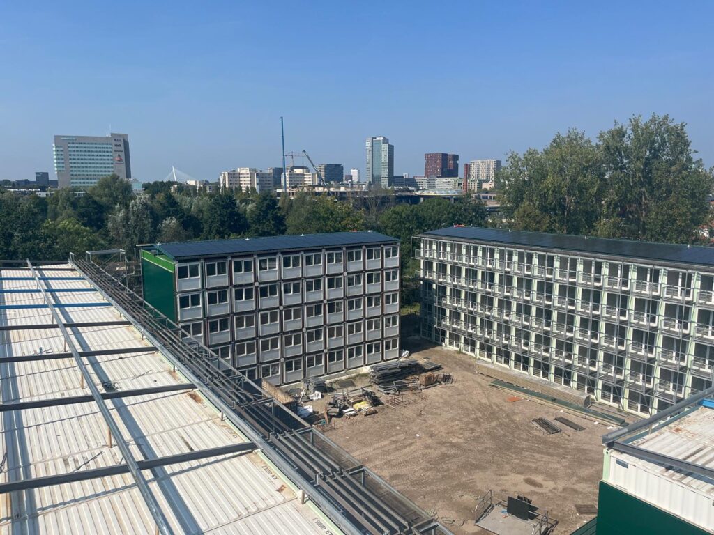 Projecten: groene en witte containerwoningen rondom een binnenplaats, met stedelijke hoogbouw zichtbaar op de achtergrond.